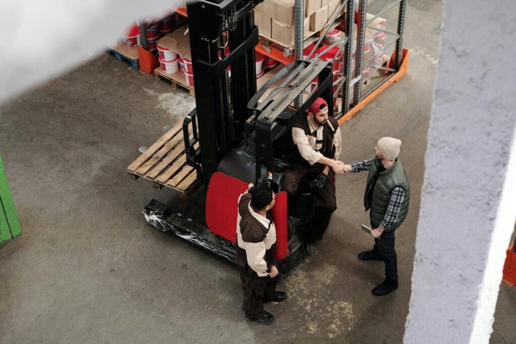 Three warehouse workers in conversation and handshake next to a forklift in an industrial setting.